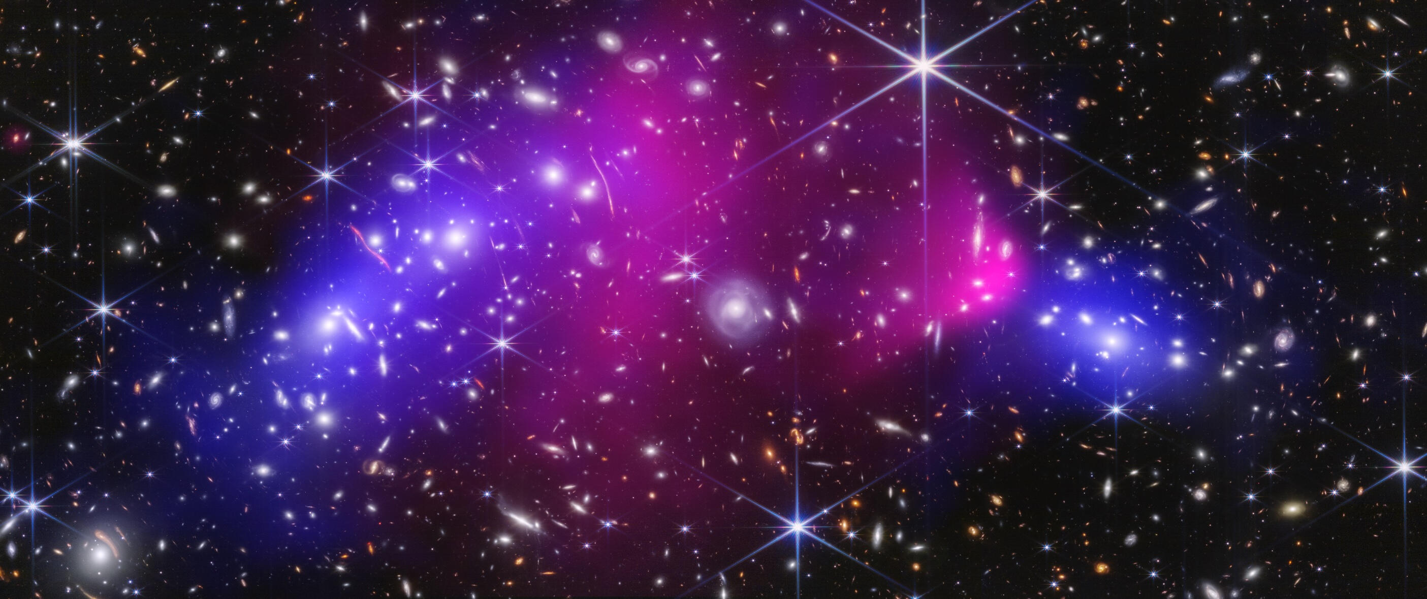 A multi-wavelength composite image of the Bullet Cluster (1E 0657-56) showing two colliding galaxy clusters. A dense field of distant, colorful galaxies captured by James Webb’s NIRCam is overlaid with glowing pink clouds representing hot X-ray gas from th